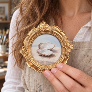 Op de afbeelding: Een klein rond schilderij van een geopende oesterschelp in een sierlijke, goudkleurige lijst. Het kunstwerk heeft een textuurrijk canvasoppervlak met de schelp tegen een lichte, neutrale achtergrond, gevat in een decoratieve, reliëfrijke rand.