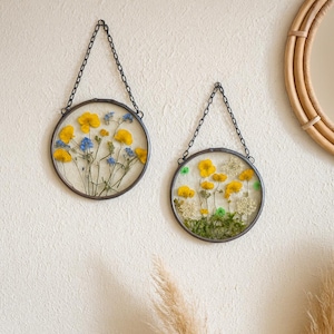May include: Two circular glass frames with dried flowers, suspended by chains. One contains yellow and blue flowers, the other yellow and white. The frames are a decorative botanical wall art piece, adding a touch of nature.