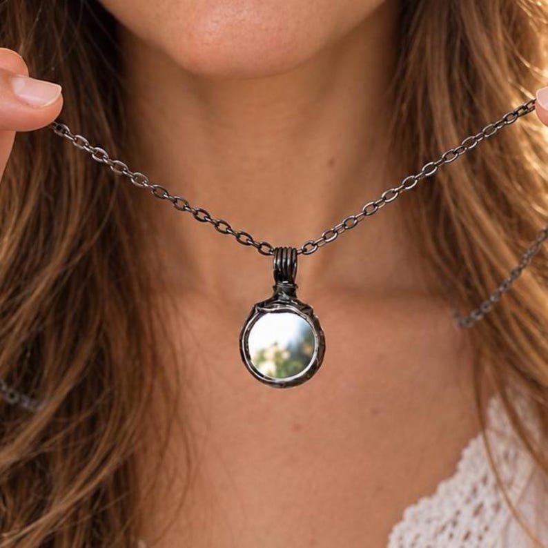 May include: A dark gunmetal-toned necklace featuring a circular pendant with a reflective mirror-like center. The pendant is suspended from a matching dark metal chain with oval links, showcasing a smooth, polished finish throughout.