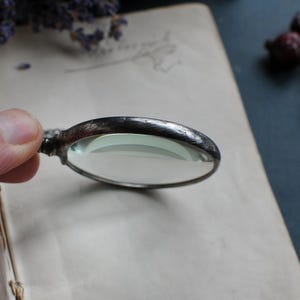 May include: A vintage magnifying glass with a silver-toned metal frame and a clear, oval lens. The handle is intricately designed. The magnifying glass is resting on an open book with aged, yellowed pages, suggesting a focus on antique or historical items.