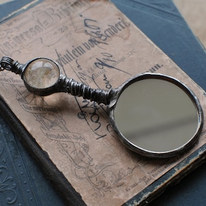 May include: Antique silver-toned magnifying glass with a round mirror and a decorative handle. The handle has a textured design and a small, round, clear stone at the top. The magnifying glass rests on an old book with German text.