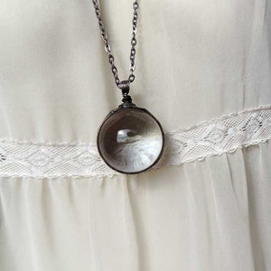 May include: A silver chain necklace with a round, clear glass pendant. The pendant is slightly convex and has a reflective surface.