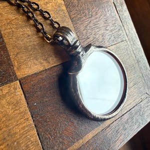 May include: A circular pendant with a textured, dark metallic finish and a smooth, reflective white center, suspended from a matching dark metal chain. The pendant features a prominent, ridged bail at the top where it connects to the chain links.