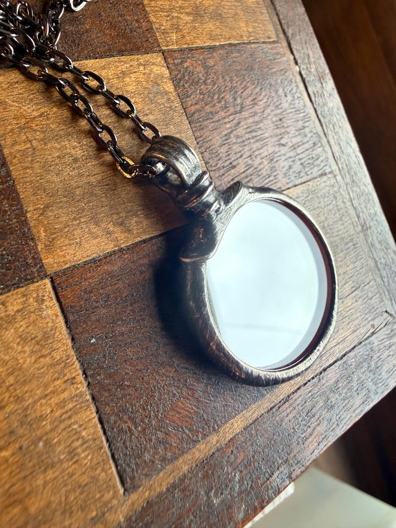 May include: A circular pendant with a textured, dark metallic finish and a smooth, reflective white center, suspended from a matching dark metal chain. The pendant features a prominent, ridged bail at the top where it connects to the chain links.