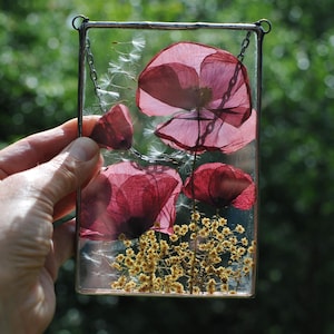 Op de afbeelding: Een rechthoekig glazen kunstwerk met geperste rode klaprozen en gele gedroogde bloemen. De bloemen zijn ingekapseld in heldere hars, met een zilveren ketting om op te hangen. De achtergrond is een vage groene kleur.