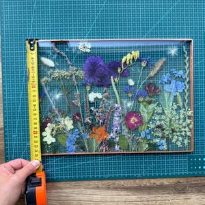 Op de afbeelding: Een rechthoekige glazen lijst met een koperkleurige metalen rand met een arrangement van geperste gedroogde bloemen in paarse, blauwe, groene en oranje tinten. Een geel meetlint toont dat de hoogte van de lijst ongeveer 25 cm is.
