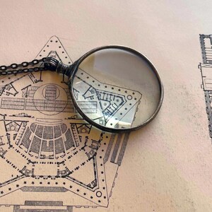 K&ouml;nnte beinhalten: Eine silberne Lupe mit einer runden Linse liegt auf einer alten Architekturzeichnung eines Geb&auml;udes mit einem sternf&ouml;rmigen Grundriss.