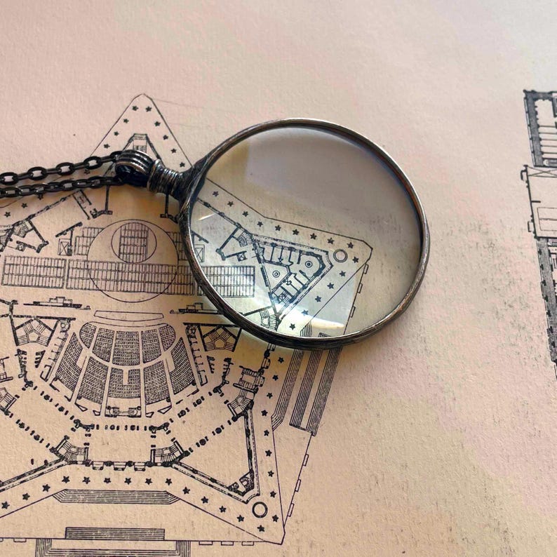 K&ouml;nnte beinhalten: Eine silberne Lupe mit einer runden Linse liegt auf einer alten Architekturzeichnung eines Geb&auml;udes mit einem sternf&ouml;rmigen Grundriss.