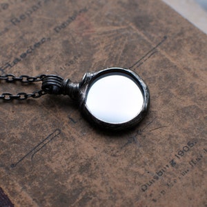 May include: A circular pendant necklace featuring a polished, reflective mirror surface encased in a dark, textured, oxidized metal frame with a matching dark metal chain link necklace attached to the top bail.