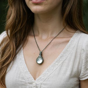 May include: A silver-toned pendant necklace featuring a circular, moon-like design. The pendant hangs from a delicate chain. The necklace is worn over a white, button-down shirt.