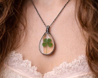 Può includere: Una collana argentata con un ciondolo a forma di goccia. Il ciondolo racchiude un quadrifoglio conservato in resina trasparente. La collana è indossata su un top di pizzo color crema.