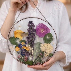 May include: A circular glass wall hanging featuring pressed dried flowers in shades of purple, yellow, and green. The arrangement includes a large pink rose, a tall purple spike, yellow petals, and green leaves, all encased in a thin metal frame.