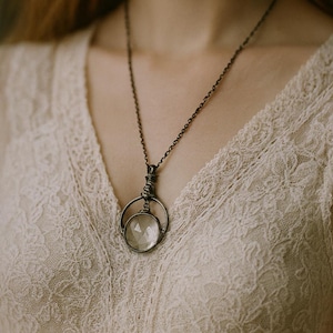 Puede incluir: Un collar con colgante de estilo vintage con una piedra clara y facetada, encerrada en un marco circular envuelto en alambre. El collar tiene un acabado oscuro y antiguo y cuelga de una delicada cadena. El colgante destaca sobre una prenda de encaje color crema.