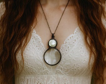 May include: A vintage-style magnifying glass pendant necklace. The pendant features a circular magnifying glass below a smaller, round, pearlescent accent. The chain and frame have a dark, antique finish. The necklace is worn over a white lace dress.
