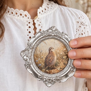 Può includere: Una piccola cornice circolare color argento con dettagli floreali ornamentali che circonda un'immagine stampata di una quaglia marrone e marrone chiaro in un campo di erba alta e piccoli fiori blu. La cornice ha una finitura metallica.