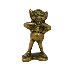 Peut inclure: Petite figurine en laiton représentant un gobelin souriant, les mains sur les hanches. Le gobelin a de grandes oreilles et un ventre rond, debout sur un petit socle. La figurine a un aspect vintage, sur fond blanc.