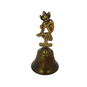 May include: A vintage brass bell featuring a seated pixie figurine. The bell has a flared shape and a clapper. The word "DEVON" is inscribed on a small plaque. This decorative bell is likely a souvenir, possibly from Devon, England.