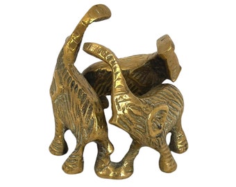 May include: A brass figurine featuring three elephants standing in a circle with their trunks raised upward. The metal surface has a textured, etched finish with visible lines detailing the skin and ears of each elephant figure.