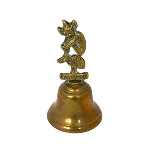 May include: A brass bell featuring a seated imp figurine. The bell has a flared base and a clapper. The word "Cornwall" is inscribed on a small plaque below the imp. The bell is a warm gold colour, typical of brass.