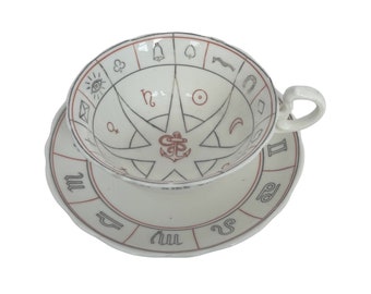 May include: A white ceramic teacup and saucer set featuring red and black printed symbols. The interior of the cup displays a central anchor, a compass star, and various astrological icons. The saucer rim is decorated with zodiac signs.