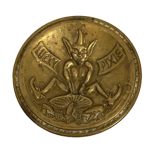 May include: A vintage brass button with a detailed relief design. The central image features a seated pixie figure atop a mushroom, with the words "LUCKY PIXIE" arched around the top. The button has a detailed edge.