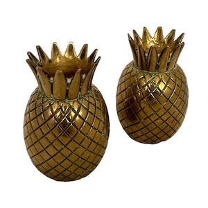 May include: Two gold pineapple-shaped candle holders. The candle holders are made of metal and have a detailed pineapple design.