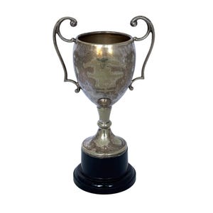 Peut inclure: Une coupe trophée argentée dotée de deux anses ornées et incurvées sur les côtés. Le corps de la coupe présente une finition métallique ternie avec un texte gravé peu visible. Elle est montée sur une base noire circulaire et lisse.