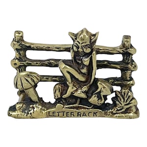 May include: A brass letter rack featuring a seated fairy figure in front of a rustic fence. The fairy is perched on a mushroom, with the words "LETTER RACK" etched on the base. The rack has a detailed, antique appearance.