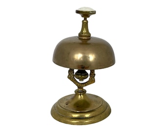 May include: A vintage brass bell with a domed top and a decorative base. The bell has a small white knob on top and ornate detailing on the handle. The bell is likely used for calling attention or signaling.