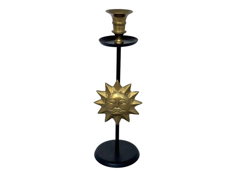 Peut inclure: Un bougeoir d&eacute;coratif en laiton avec un motif de visage de soleil. Le bougeoir a une base et une tige noires, avec un soleil en laiton au centre. Le haut contient un porte-bougie.