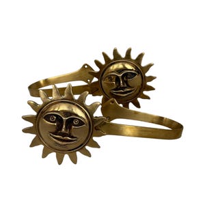 May include: Two brass-colored curtain tiebacks. Each tieback features a sun face with a smiling expression and radiating sun rays. The tiebacks are designed to hold curtains and add a decorative touch to any room.