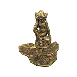 May include: A brass figurine of a seated fairy with pointed ears, resting on a leaf-shaped base. The fairy is holding its head with one hand. The base has detailed leaf vein patterns. The figurine is made of brass.