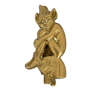 May include: A brass door knocker depicting a seated goblin with a playful expression. The goblin has pointed ears, a skirt, and clasped hands. The knocker is set against a white backdrop, showcasing its intricate details.