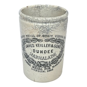 May include: Antique white ceramic marmalade jar with a cracked glaze. The jar features black text that reads "JAMES KEILLER & SONS DUNDEE MARMALADE" and "GRAND MEDAL OF MERIT VIENNA 1873" and "PRIZE MEDAL FOR LONDON, 1862".