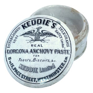 May include: A vintage white ceramic jar with a lid, marked with "KEDDIE'S" and "GORGONA ANCHOVY PASTE". The lid shows an eagle emblem and the text "ESTABLISHED 1851". The jar is open, showing the inside.