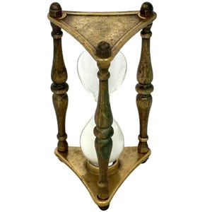 May include: An hourglass featuring a triangular brass frame with a weathered, antique finish. The structure consists of three vertical, turned pillars connecting a top and bottom triangular plate. A clear glass bulb sits in the centre.