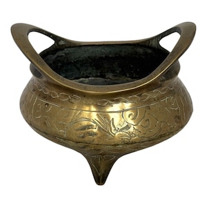 May include: A brass bowl with a rounded body, three short legs, and two arched handles extending from the rim. The exterior surface features engraved line patterns and floral motifs, while the interior has a dark, weathered patina finish.