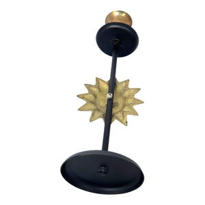 Peut inclure: Un bougeoir en m&eacute;tal noir avec un motif de soleil dor&eacute;. Le bougeoir a une base ronde et un petit sommet rond pour tenir une bougie. Le soleil est centr&eacute; sur la tige.
