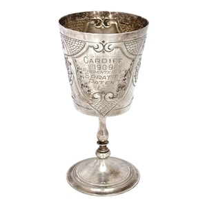 May include: A silver-toned metal trophy cup featuring an engraved shield design with the text CARDIFF 1909 PRESENTED BY SPRATTS PATENT LTD. The cup has a flared rim, a slender stem with a central node, and a circular base with a smooth, polished finish.
