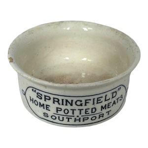 May include: Off-white ceramic bowl with a slightly flared rim. The bowl has a dark blue oval label with the text "SPRINGFIELD HOME POTTED MEATS SOUTHPORT". The bowl has some discoloration and wear.