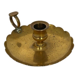 May include: A brass candlestick holder with a scalloped edge and a handle. The holder has a circular base and a cup-shaped top for a candle. The brass has a golden hue and shows signs of age. The design is vintage.