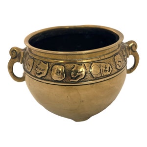 May include: A brass-coloured bowl with a dark interior and two handles. The bowl is decorated with a band of embossed theatrical masks. The masks are of various expressions. The bowl has a rounded shape.