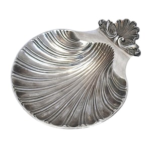 May include: A silver-toned metal dish shaped like a scallop shell. The dish features a concave interior with radiating fluted grooves and a decorative, ornate handle at the top edge that resembles a fan-shaped shell motif with scrollwork details.