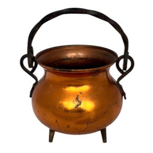 May include: A small, copper-colored cauldron with a dark, wrought iron handle and three short legs. The cauldron has a rounded body and a wide opening, with a slightly textured surface. The handle is arched and attached to the sides.