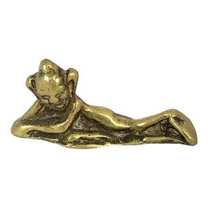 May include: A small, golden figurine depicting a reclining figure. The figure is lying down with one arm supporting the head. The figurine has a smooth, polished finish and is likely made of brass or a similar metal.