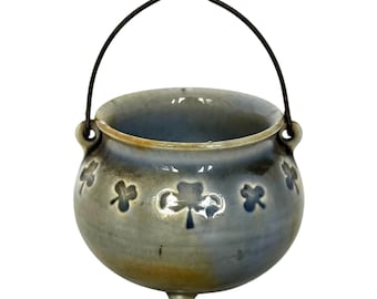 May include: A round ceramic pot with a glossy blue and tan glaze, featuring embossed shamrock patterns around the exterior. The pot has three small feet at the base and a thin, dark metal wire handle attached to two side loops for carrying.