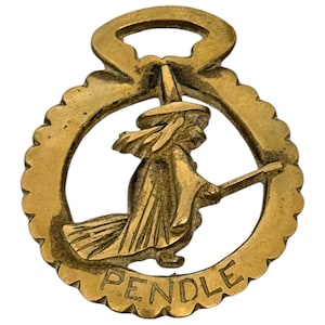 May include: A gold-colored brass horse brass with a cutout design of a witch riding a broomstick. The brass has a scalloped edge and the words "PENDLE" are inscribed at the bottom.