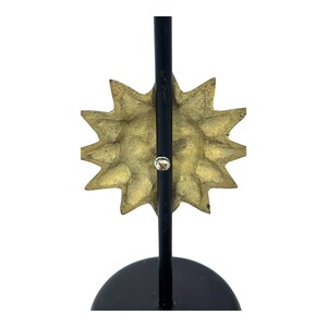 Peut inclure: Une sculpture d&eacute;corative en forme d'&eacute;clat de soleil avec un visage textur&eacute; de couleur or et des rayons pointus. L'&eacute;clat de soleil est mont&eacute; sur un support en m&eacute;tal noir avec une base circulaire.