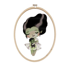 May include: Cross-stitch embroidery hoop featuring a green-skinned cartoon monster girl in a white dress holding a smaller green monster doll.