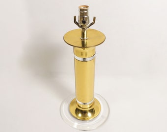 May include: A gold table lamp with a clear acrylic base. The lamp has a cylindrical shade and a simple design.